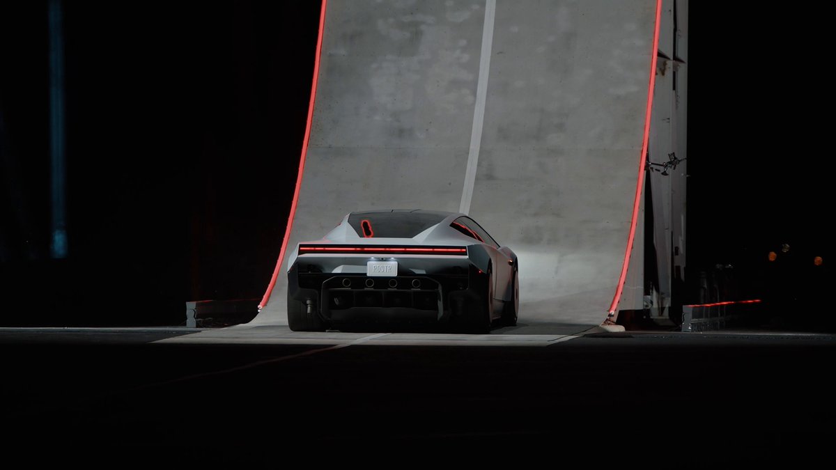 Rear view of the redesigned Tesla Roadster with an Optimus robot in the cabin approaching a vertical C-shaped stunt ramp at night