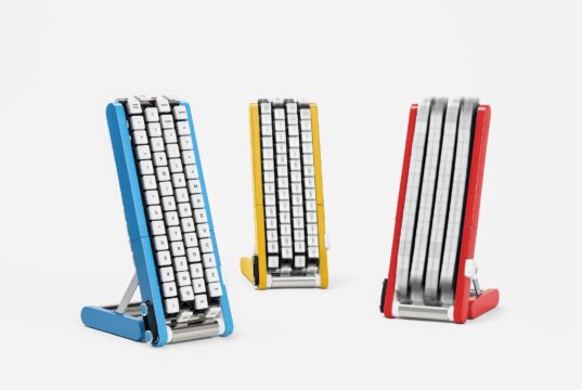 Three conveyor-belt keyboards on stands in blue, yellow and red, each with white keycaps mounted on four vertical belts; the red unit's belts are blurred with the keys in motion