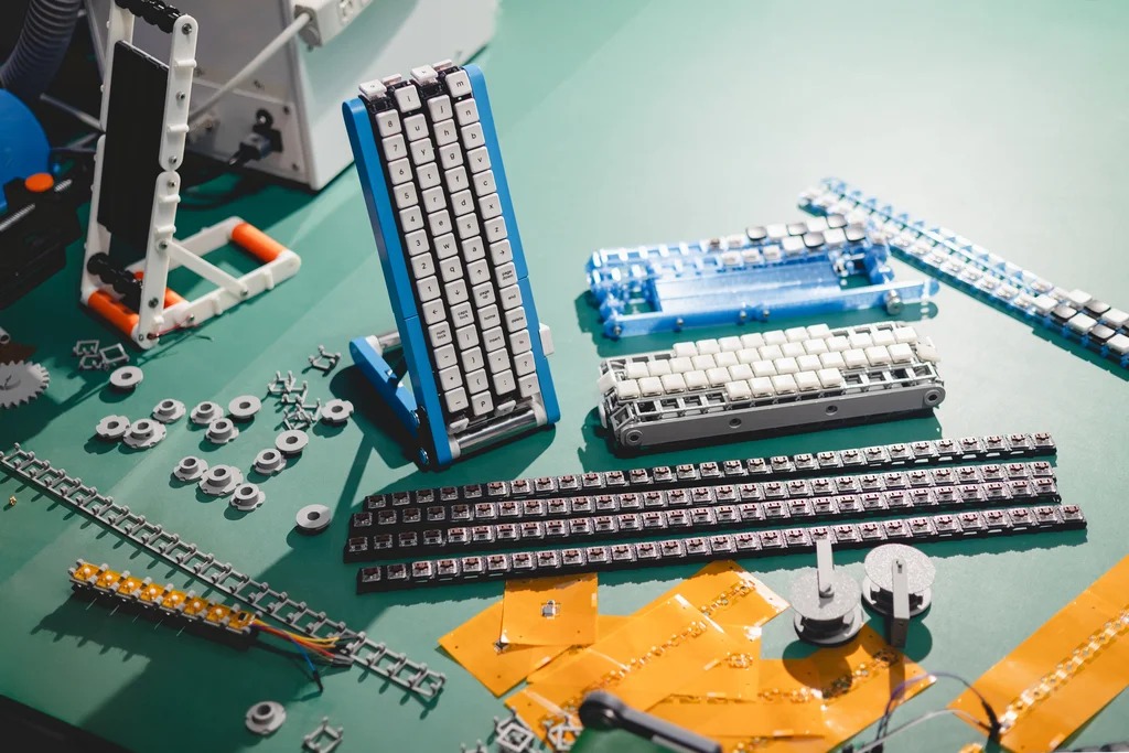 Workbench photo of the conveyor-belt keyboard being built: the assembled blue unit standing beside loose keycap strips, white rollers, orange flex circuits, screws and 3D-printed parts on a green mat