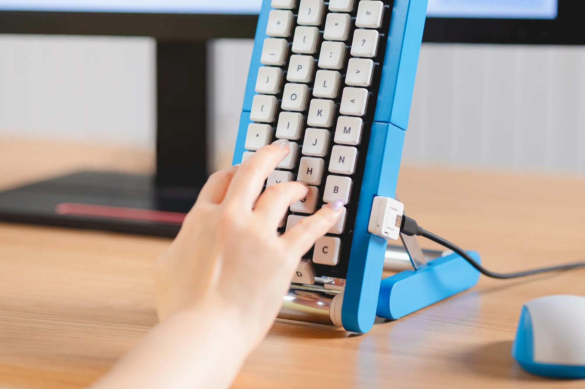 A single left hand typing on the blue conveyor-belt keyboard at a wooden desk, a white and blue mouse resting on the desk beside it
