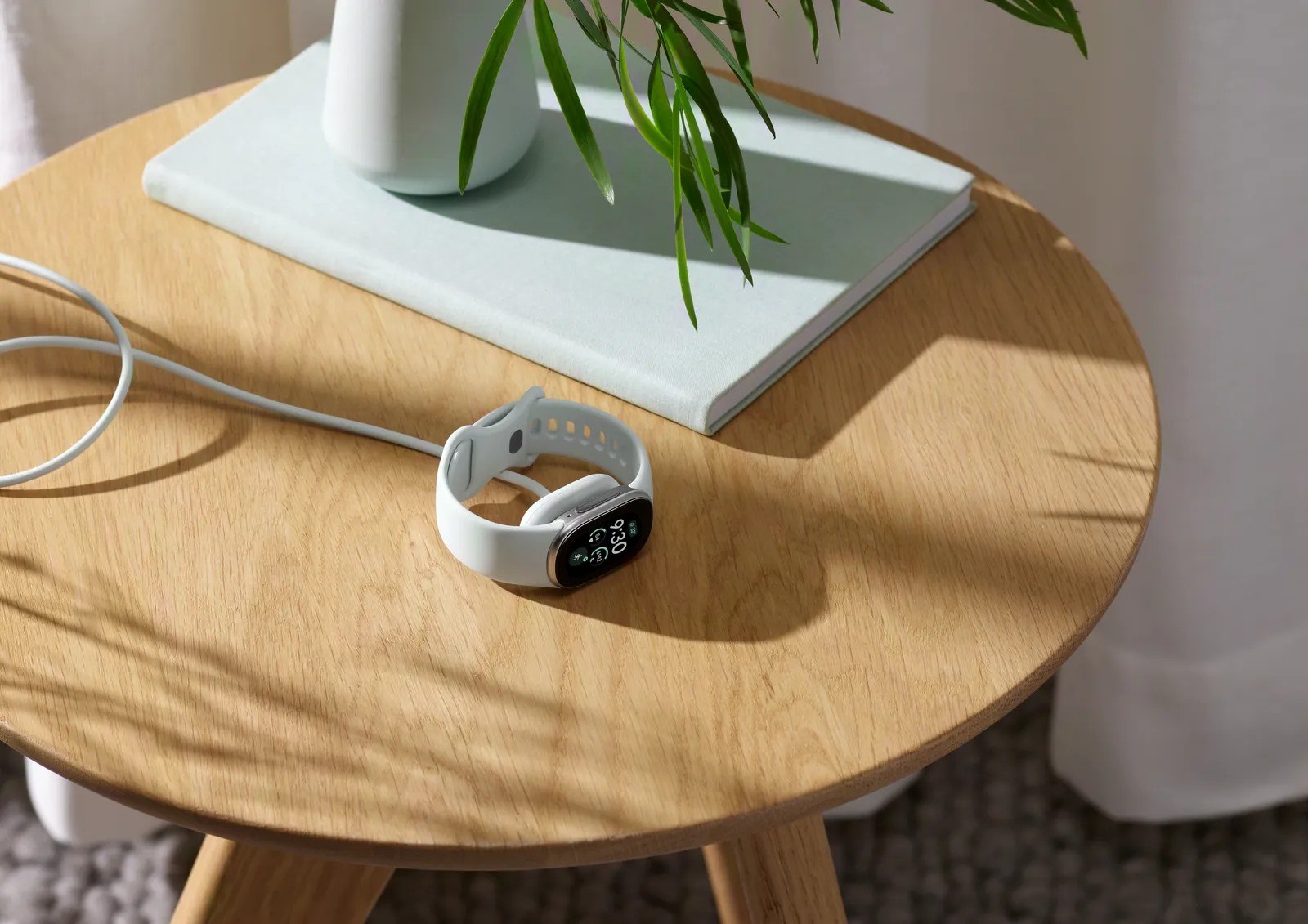 A Fitbit Edge with a light band resting on a round wooden table beside a book and its charging cable
