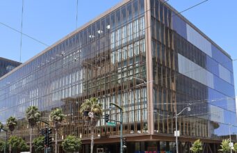 OpenAI Engineer Charged Over Hidden Recording Device in Executive Room Glass-fronted office building at 1515 Third Street in San Francisco's Mission Bay, with palm trees and a traffic signal at the corner