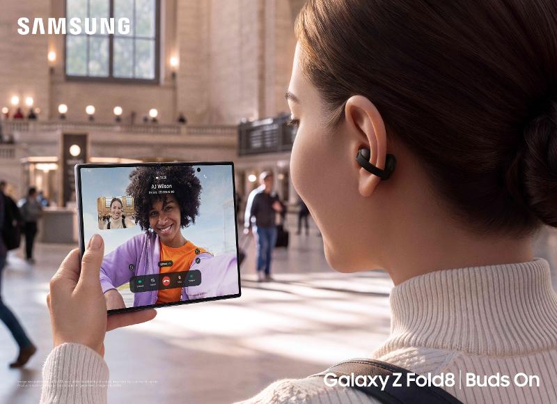 A person wearing a black clip-on Galaxy Buds On earbud holds an open foldable phone showing a video call in a station concourse