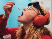 Skullcandy Stays Separate After Bose Buys the Brand A person in a red shirt and orange headphones holds a dropper of pink liquid near an open mouth, with black goggles on their forehead