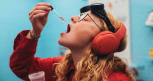 Skullcandy Stays Separate After Bose Buys the Brand A person in a red shirt and orange headphones holds a dropper of pink liquid near an open mouth, with black goggles on their forehead