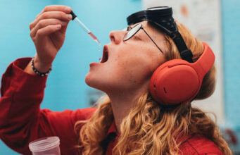 Skullcandy Stays Separate After Bose Buys the Brand A person in a red shirt and orange headphones holds a dropper of pink liquid near an open mouth, with black goggles on their forehead