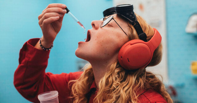tmm-skullcandy-site-image A person in a red shirt and orange headphones holds a dropper of pink liquid near an open mouth, with black goggles on their forehead