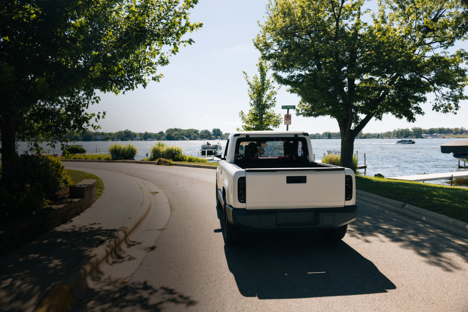 White pickup truck seen from behind on a curved lakeside street, with two occupants in the cab and an open cargo bed
