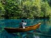A person seated in a wooden canoe on a clear river, filming a dense jungle shoreline with a smartphone