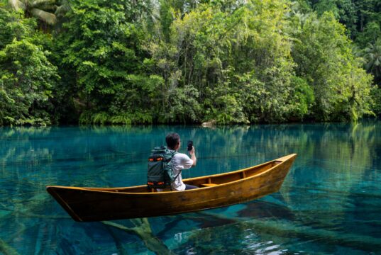 A person seated in a wooden canoe on a clear river, filming a dense jungle shoreline with a smartphone