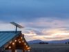 Starlink dish mounted on a green metal roof with string lights at dusk, fields and mountains behind it