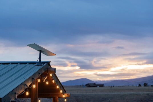 Starlink dish mounted on a green metal roof with string lights at dusk, fields and mountains behind it