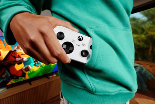 A person in a teal hoodie pulls a white Xbox Nano Controller from a front pocket, with a television on in the background