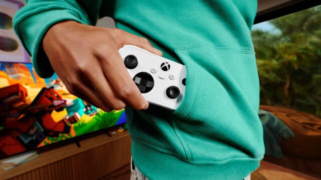 tmm-xbox-nano-controller-hero A person in a teal hoodie pulls a white Xbox Nano Controller from a front pocket, with a television on in the background